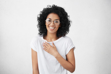 Wall Mural - Happy beautiful warm-hearted young dark-skinned female wearing big round spectacles and white t-shirt smiling and holding hand on her breast while watching touching romantic scene in TV series