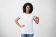 © wayhome.studio  - Clothing, design and advertising concept. Indoor shot of positive friendly young mixed race female in glasses pointing at copy space on her blank white t-shirt for your text or promotional content