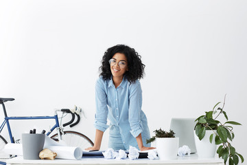 Cheerful young dark-skinned female interior designer wearing stylish glasses and blue shirt standing at her workplace, placing hands on desk after finished working on new project, smiling happily