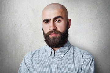 A confident bald male with thick black eyebrows and beard wearing checked shirt having gloomy expression posing against white background.People, fashion, lifestyle concept.