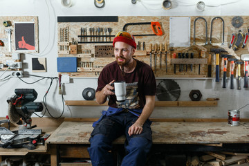 A young professional carpenter with a beard sits in the workshop and drinks coffee during the break