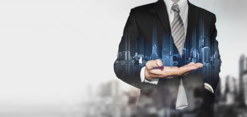 panoramic businessman holding blue hologram of modern buildings, with blur background and copy space