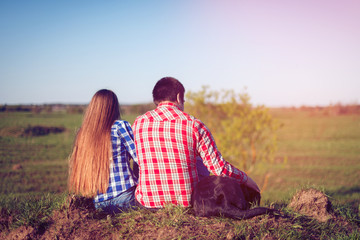 Young couple in plaid shirts and shorts and the dog is sit back and admire the scenery. Tourists rest on the hill in front of the grass field. Toned