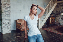 Attractive Blonde Woman In A White T-shirt Sits At Home On A Chair.