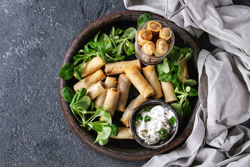 Wall Mural - Fried spring rolls with white yogurt sauces, served in terracotta plate and fry basket with fresh green salad over black texture background. Flat lay, space. Asian food