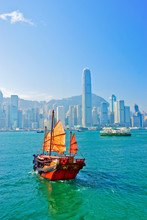 View Of Hong Kong Skyline With A Red Chinese Sailboat Passing On The Victoria Harbor In A Sunny Day.