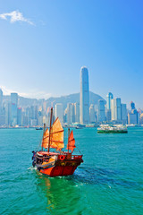 View of Hong Kong skyline with a red Chinese sailboat passing on the Victoria Harbor in a sunny day.