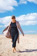 © raisondtre - Portrait of a beautiful sad business woman in a black dress and elegant coat walking barefoot on the sandy beach with his head down, on a Sunny day on blurred background of blue cloudy sky.