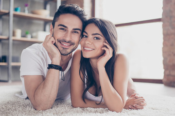 True love. Happy beautiful married couple is chilling out lying on the carpet at the floor at home indoors, looking at the camera and smiling