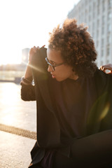 Stylish African American curly girl sitting on the roof putting hood on with sunset backlight over cityscape. Woman in glasses in early morning over urban background on the sunrise