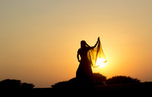 Young Woman  In Indian Clothes Dances At Sunset In The Desert On The Dunes Against Low Bushes, Sun And Sky. Rajasthan, India.