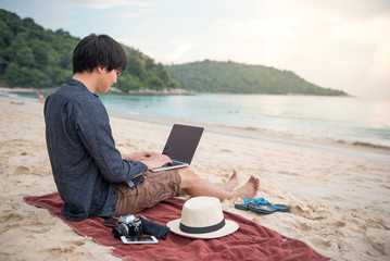 Young Asian man working with laptop computer on tropical beach, digital nomad lifestyle or freelance job concepts