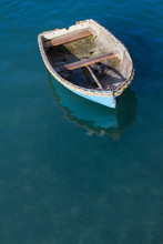 Looking Down From Above Onto A Small, Blue Rowing Boat That Has Broken Away From It's Moorings And Is Floating Away On A Calm Ocean.