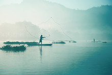 Fisherman Casting Out His Fishing Net In The River By Throwing It High Up Into The Air Early In The Blue Colored Morning To Catch Fish With His Little Fishing Boat.