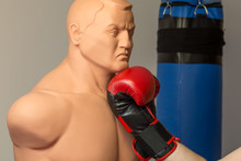 Red Boxing Gloves Punching An Uppercut On The Chin Of A Dummy Mannequin Doll On A Gym With A Blue Punching Bag On The Background