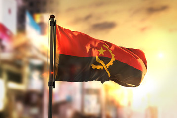 Sticker - Angola Flag Against City Blurred Background At Sunrise Backlight