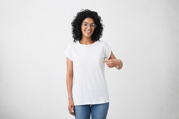 T-shirt design and advertising concept. Style and fashion. Indoor shot of cheerful smiling young African American woman with curly hair pointing index finger at copy space on her blank white top