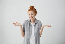 Indoor Shot Of Beautiful Caucasian Redhead 20 Y.o. Girl With Hair Knot Shrugging Her Shoulders And Smiling Broadly Feeling Confused While Refusing To Go Out On Date With Guy She Doesn't Like