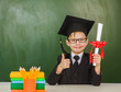 © Ermolaev Alexandr - Happy boy with diploma in graduation cap showing thumbs up near a school board