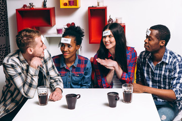 Young international company play funny game who I am. Four smiling people sit at the table with paper stickers. Home leisure, party, laugh, imagination concept.