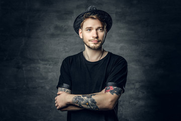 Handsome male with crossed tattooed arms, dressed in a black t shirt and tweed hat.