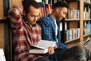 Wall Mural - Young concentrated two african men reading books using laptop