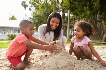 Teacher At Montessori School Playing With Children In Sand Pit