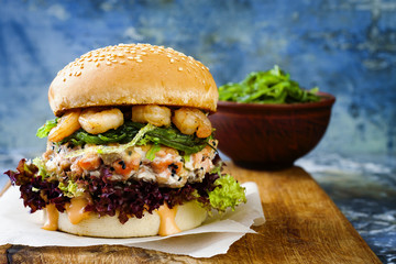 Wall Mural - Asian style salmon burger with grilled shrimps, seaweed, lettuce and spicy sriracha mayo sauce served on pieces of brown paper on a rustic wooden board. Bright blue background with copyspace