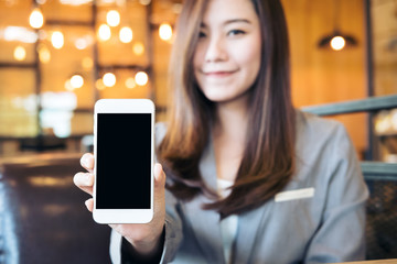 Wall Mural - Mockup image of an Asian beautiful business woman holding and showing white mobile phone with blank black screen and smiley face in cafe