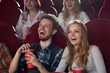 Positive couple of two, boyfriend in checked shirt and girlfriend in gray blouse surprised smiling, watching comedy movie together, holding red cup with cola. Best friends having fun at cinema hall.