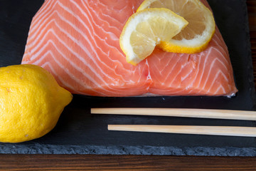 Wall Mural - Fresh salmon fillet with lemon and wooden chopsticks on dark stone background. Top view. Closeup