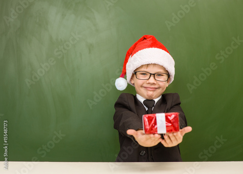 Fényképezés Happy boy in red christmas hat with gift near empty green blackboard