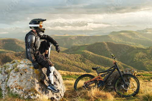 Fotomural A professional downhill rider sits on a stone resting after a tense race on a mt