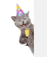 Happy cat in birthday hat holding glass of champagne and peeking from behind empty board. isolated on white background