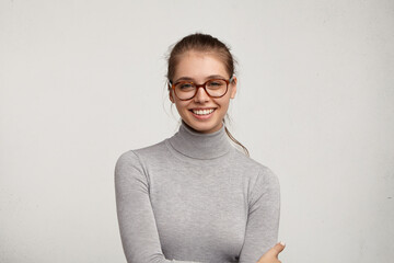 Headshot of cute student girl wearing grey turtleneck sweater and stylish eyeglasses smiling broadly, having carefree and relaxed look while resting at home after morning lectures at college