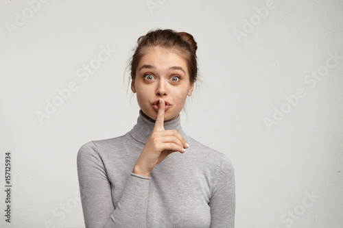 Fotografie, Obraz Studio shot of wide eyed pretty girl in turtleneck asking for silence or secrecy, holding finger at lips