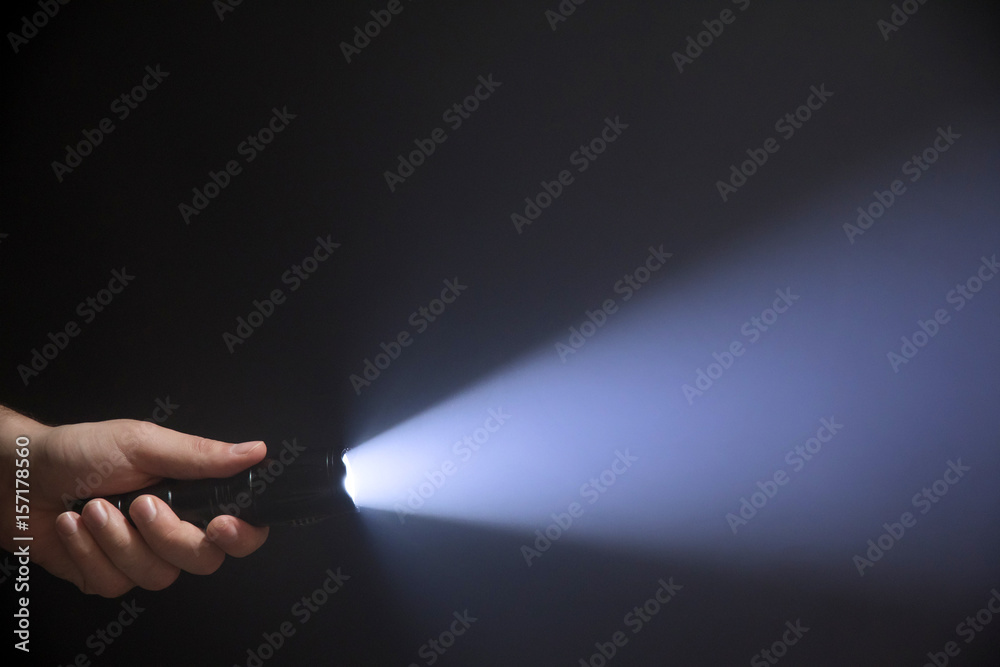 Black flashlight with wide beam in male's hand isolated from left side of the frame on black background - obrazy, fototapety, plakaty 