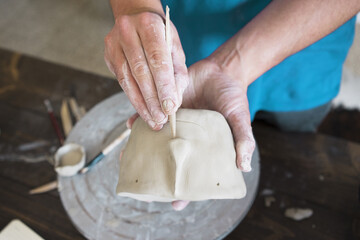 Clay stained hands of sculptor, close up view. Man in blue t-shirt creates style of ceramic pot with funny face using wooden tool in his home white studio with wooden dark brown table.