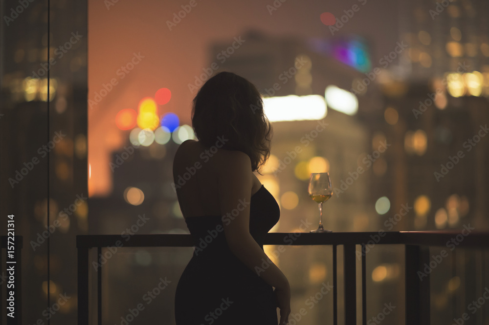 Beautiful young woman on the balcony in a black dress with a glass of ...