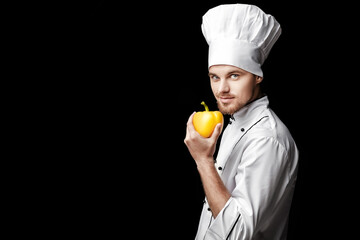 Wall Mural - Young bearded man chef In white uniform holds yellow bell pepper on  black background