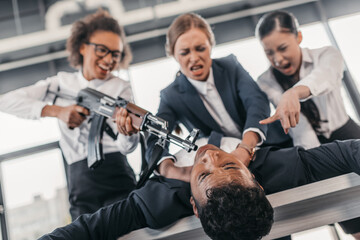 Three young angry businesswomen with rifle punishing businessman lying on table, business team meeting concept