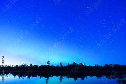 Early night sky with trees silhouetted and reflecting in calm waters, stars and northern lights just beginning