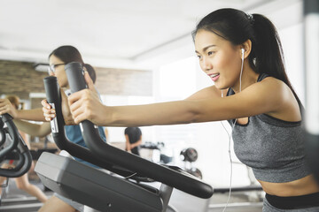 Asian woman with earphones on exercise bike at the gym.