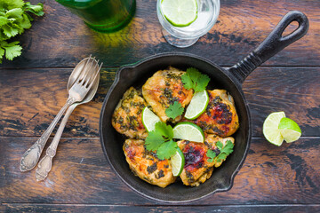 Spicy and crispy homemade cilantro lime chicken thighs in a black cast-iron skillet on the rustic wooden table, top view.