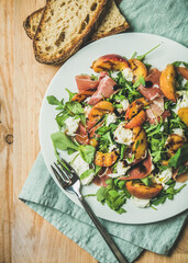 Wall Mural - Arugula, prosciutto, mozzarella and grilled peach salad in white plate over blue napkin and wooden board, top view, vertical composition