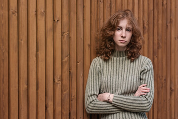 A beautiful red-haired girl in a green sweater is standing by the wooden wall. She crosses her arms and looks severely at the camera.