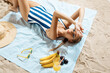 Tanned young woman taking unhealthy sunbath on a summer day on a beach hiding from the sun with straw hat and glasses. Trendy make-up smokey eyes. Drinking smoothie or orange juice with bananas and