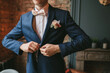© Alexandra - A young groom in a blue suit and a pink bow tie button his jacket against the backdrop of a brick wall and a fireplace