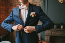A Young Groom In A Blue Suit And A Pink Bow Tie Button His Jacket Against The Backdrop Of A Brick Wall And A Fireplace