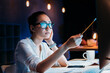 © LIGHTFIELD STUDIOS - Young businesswoman in eyeglasses holding pencil and working late in office
